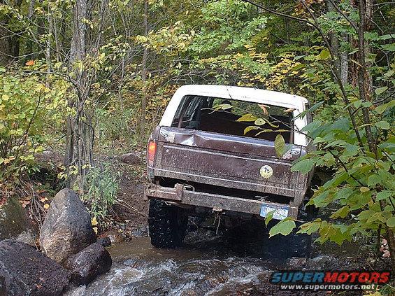 Brown and white SUV driving over rocks in a stream, trees and foliage around