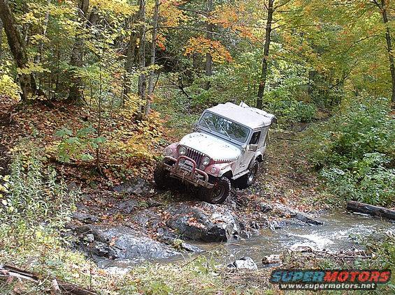 White Jeep CJ-7 off-roading through a stream in a forest, front grille and bumper visible