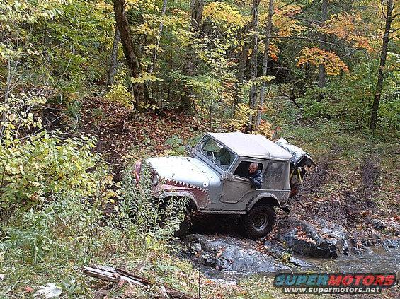 Gray Jeep CJ-5 with shark teeth painted on the hood, navigating a rocky stream