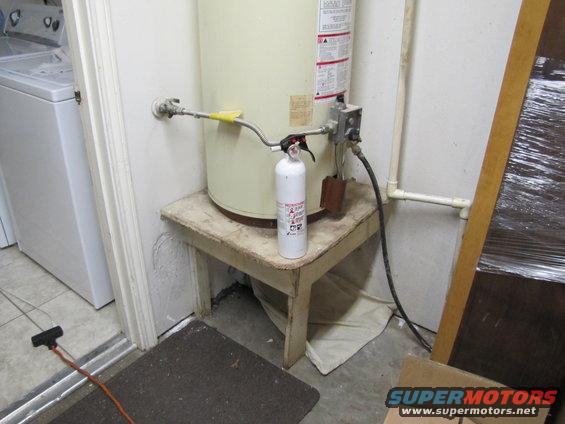 Water heater on a wooden stand with a fire extinguisher and plumbing. A washing machine is visible to the left