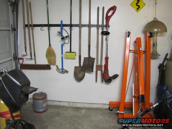 Garage wall with assorted tools hanging, including shovels, brooms, and a weed trimmer. An orange engine hoist and a welding machine are also visible