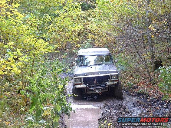 Gray Ford Bronco driving through a muddy forest trail with yellow and green foliage