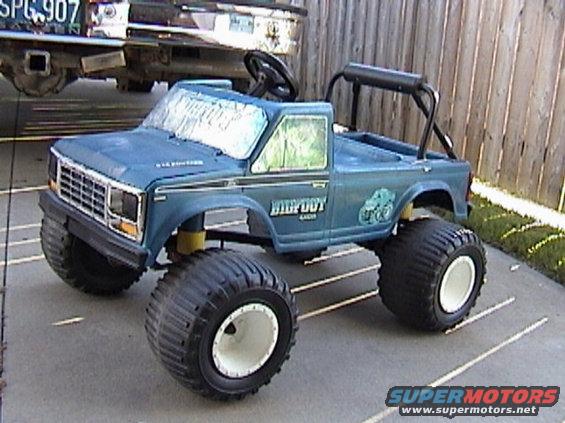 Blue toy Bigfoot monster truck with oversized tires parked on a driveway, another vehicle in background