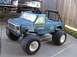 1985 Ford F-150 - A blue toy Bigfoot monster truck with large tires is parked on a driveway, with another vehicle partially visible behind it.