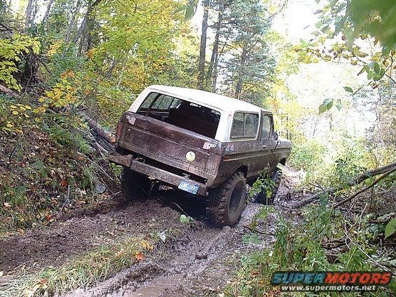 1978 Ford Bronco navigating a muddy, wooded trail with trees and fallen logs