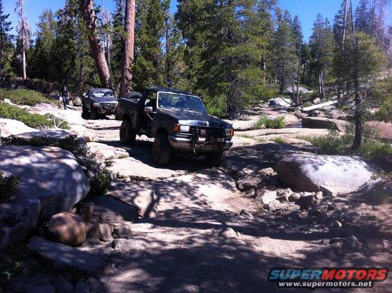 Two trucks driving on a rocky trail in a forest. One truck is in the foreground, the other is further back