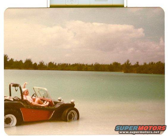 Person sits in a red and black dune buggy partially submerged in shallow water on a beach