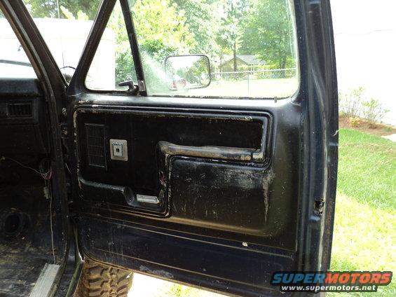 Black interior door panel with worn armrest and square vent. Visible wiring and part of a tire