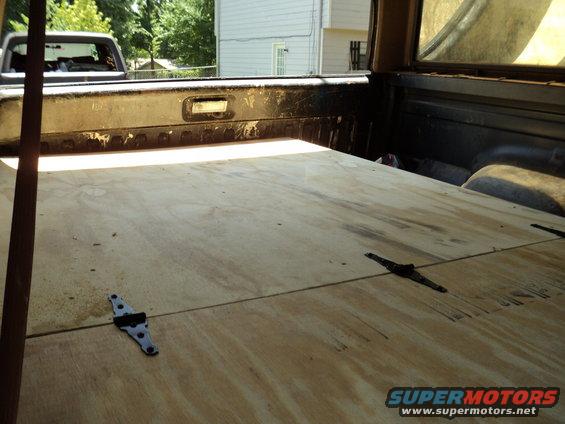 view from inside a truck bed, looking out the back window at another truck. Plywood covers the bed floor