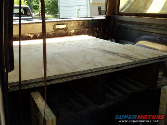 Truck bed with plywood sheets stacked on top of wooden supports. Another vehicle is visible in the background through the rear window