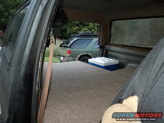 Interior view of a vehicle with a bed, cooler, and fan, looking out at a green SUV