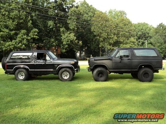 Two Ford Broncos parked on grass, one black, one matte black. The black Bronco is a 1980s model