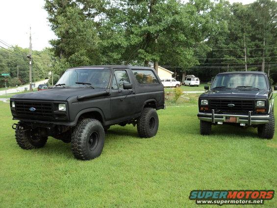 Two matte black Ford Broncos parked on a grassy lawn. The Bronco on the left is lifted with large tires