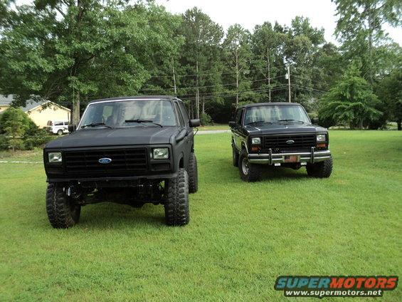 Two black Ford Broncos parked on grass. One is matte black and lifted, the other is glossy black with chrome bumpers