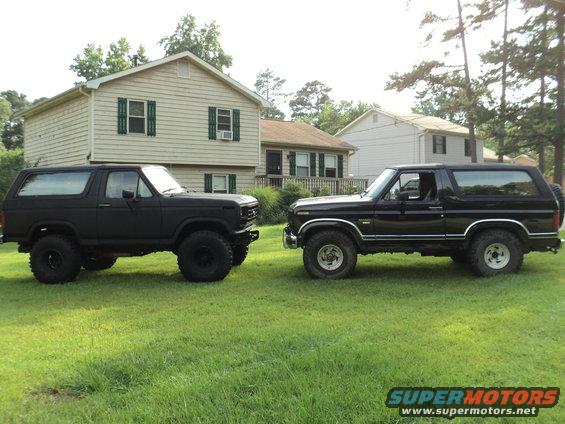 Two black Ford Broncos parked on a lawn in front of houses. The Bronco on the left is lifted with large tires