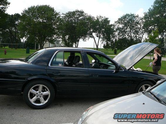 black sedan parked outside, hood up, person standing by engine. another car in foreground