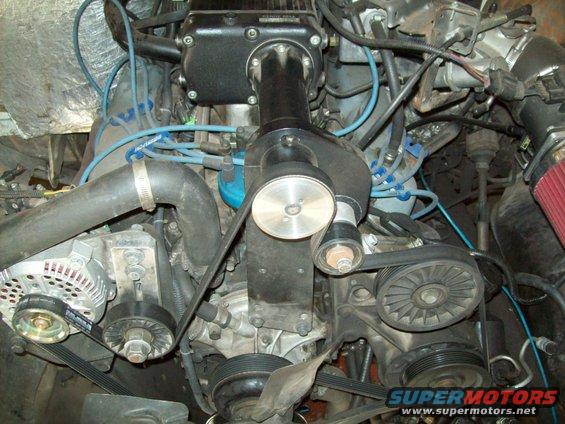 Close-up of a vehicle engine with a supercharger installed. Shows belts, pulleys, alternator, and wiring