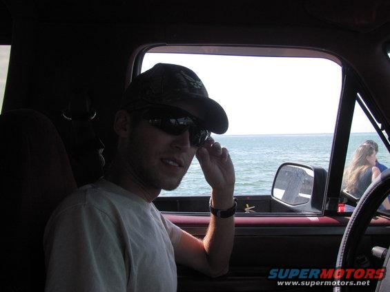 Guy in a baseball cap and sunglasses adjusts his shades inside a truck cab, looking out at the ocean. Another person is visible in the passenger seat