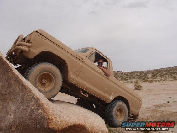 Tan 1978 Ford Bronco climbing over large rocks on an off-road trail. Driver is visible through the window