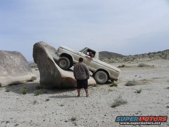 guy in a white pickup truck is driving over a big rock, another guy watches
