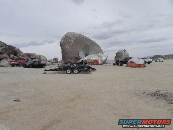 Desert scene with multiple trucks, tents, and large rock formations under a cloudy sky