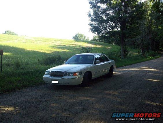 White 2000 Mercury Grand Marquis sedan parked on a gravel road, facing left, with a grassy hill and trees in the background