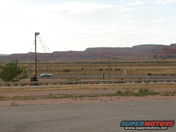 White sedan driving on highway, desert landscape with mesas behind. Pole with flags in foreground