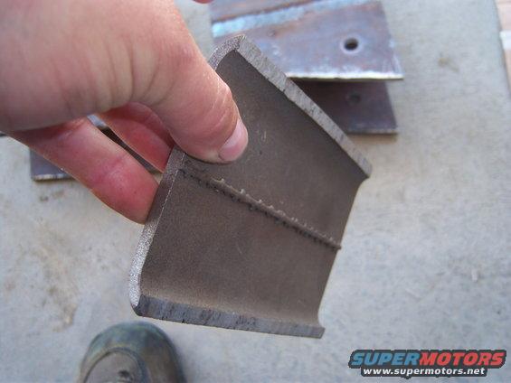 Metal bracket with welds, held by a hand. Another bracket is visible behind