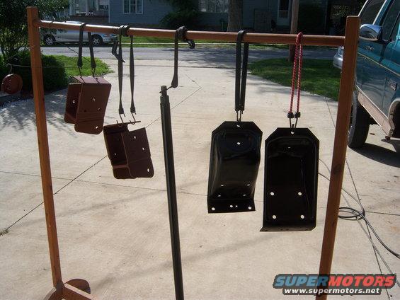 Metal parts painted brown and black hang from a wooden rack outdoors, with a teal truck visible to the right