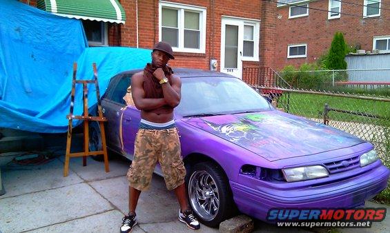 Guy in camo shorts and brown tank top stands next to a purple sedan with custom graphics on the hood. The car's front end is partially disassembled