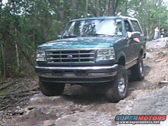 Green Ford Bronco on rocky terrain, front 3/4 view. Lifted suspension, off-road tires