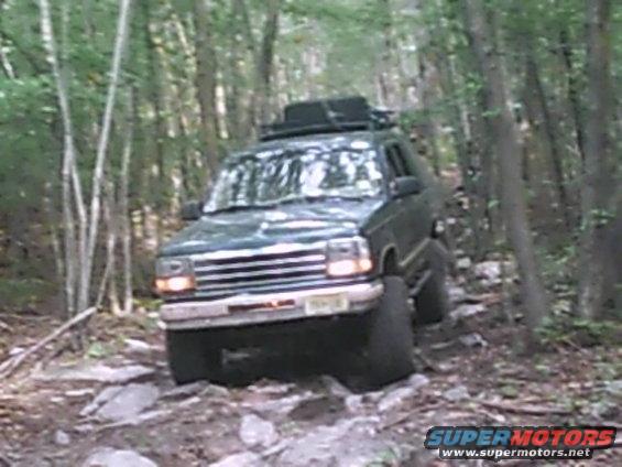 Green SUV with roof rack driving on a rocky, wooded trail. Grille suggests a 1992 model