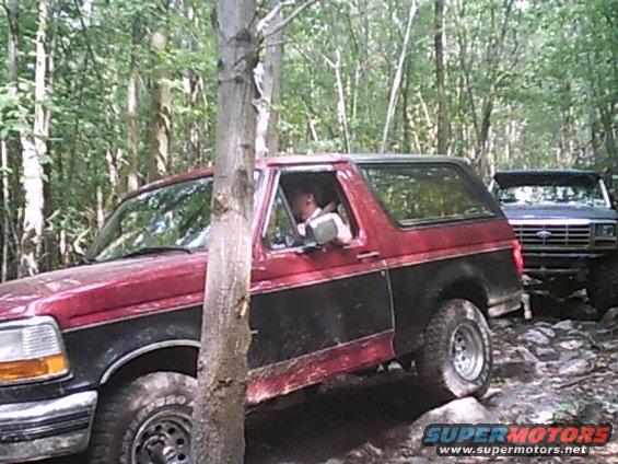 Red and black Ford Bronco driving on a rocky trail in the woods, another Bronco behind it