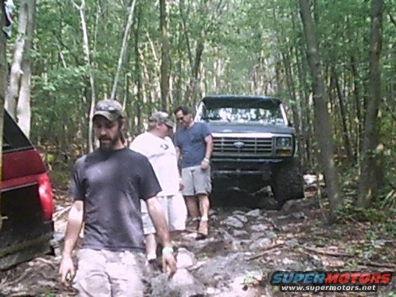 Dark blue Ford truck with a prominent grille and large tires on a rocky, wooded trail