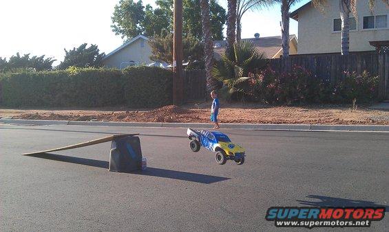 Yellow and blue remote control monster truck airborne over asphalt road, child watches