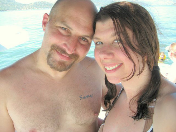 Man with goatee and woman with pigtails smiling on a boat, water and distant hills in background. Man has 'Sway' tattoo on chest