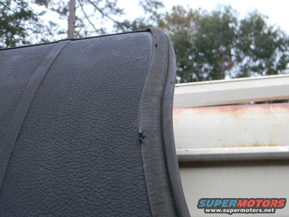 Close-up of black textured upholstery with a seam and a rubber trim piece. A white structure with rust is in the background