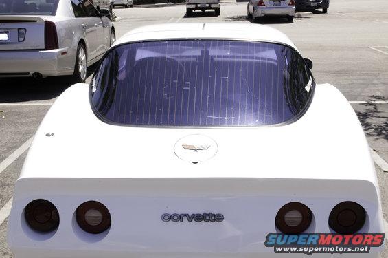 Rear of a white Corvette with 'Corvette' badging. A silver Cadillac is parked to the left
