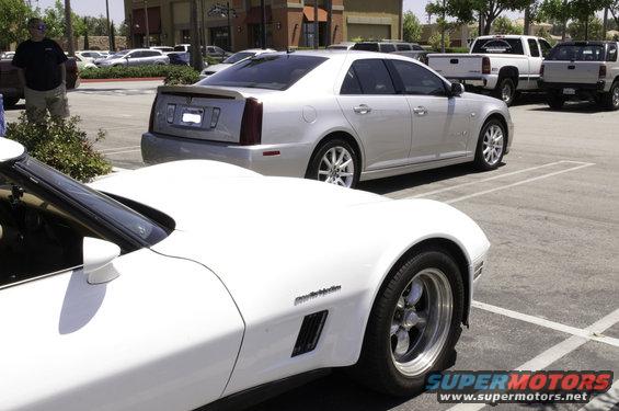 White sports car in foreground, silver Cadillac sedan behind it in parking lot. Pickup trucks visible in background