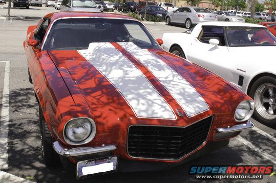 Red 1970s muscle car with dual white racing stripes on the hood and roof, parked next to a white car
