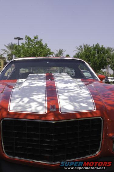 Front view of a red muscle car with a large white hood scoop and racing stripes. A small badge is on the hood