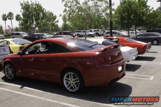 Red coupe parked in a lot, rear view. Other cars visible behind it
