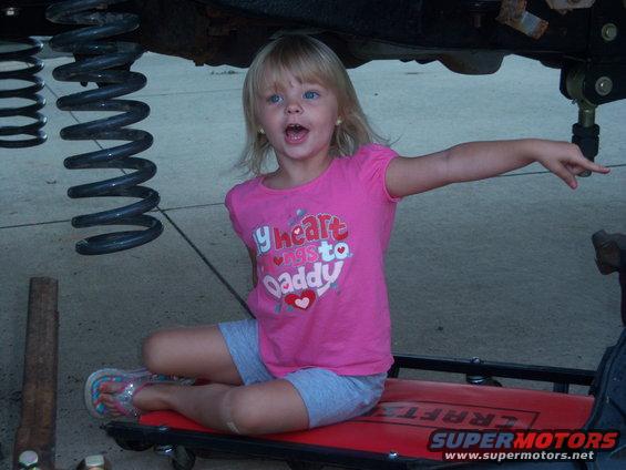 Young girl sits on creeper under vehicle, pointing