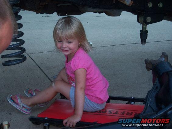 Young girl in pink shirt and gray shorts sits on a red creeper under a vehicle