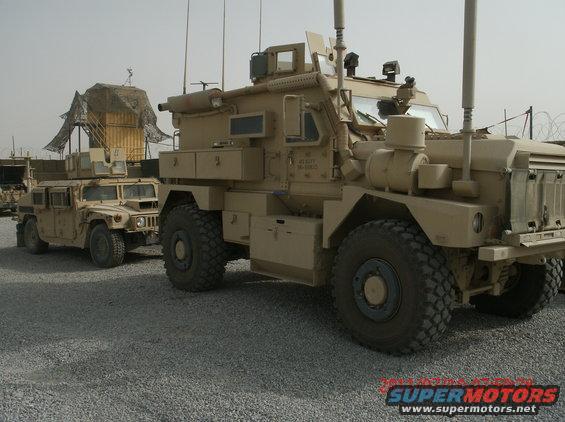 Two tan military trucks parked on a gravel lot. The truck in the foreground is larger and more heavily armored than the truck behind it