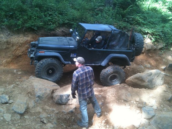 Black Jeep Wrangler on rocky terrain, front-left tire elevated, driver visible