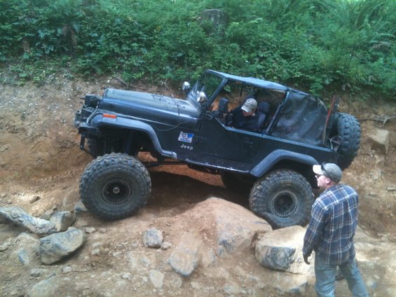 Modified Jeep Wrangler with large tires navigating a rocky off-road trail. A person is visible in the driver's seat