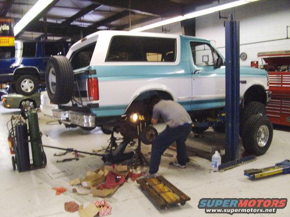 1994 Ford Bronco on a lift with a mechanic working on the rear axle, another vehicle in background