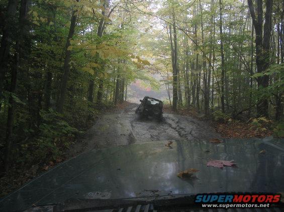Dark SUV stuck in mud on a wet forest road, viewed from another vehicle's hood