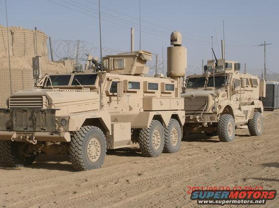 Two tan military armored vehicles parked on a dirt road with barbed wire and sandbag barriers in the background